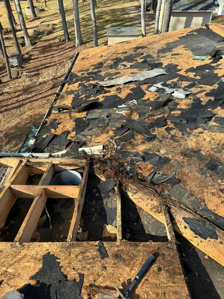 Damaged rooftop with missing shingles and exposed wooden framing amid trees and debris