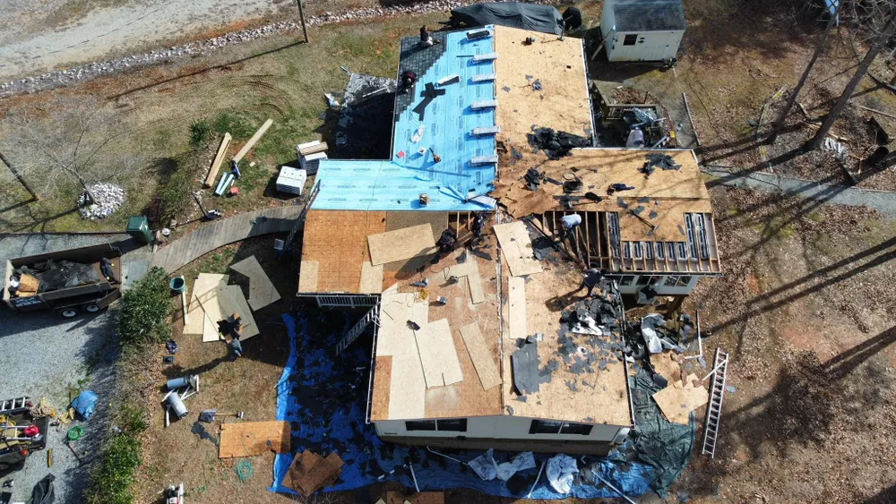 Aerial view of a storm-damaged house with a blue tarp roof and debris scattered around the yard