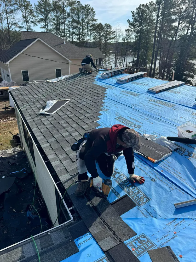 Workers installing shingles on a sloped roof under blue tarp, with a house and trees in the background