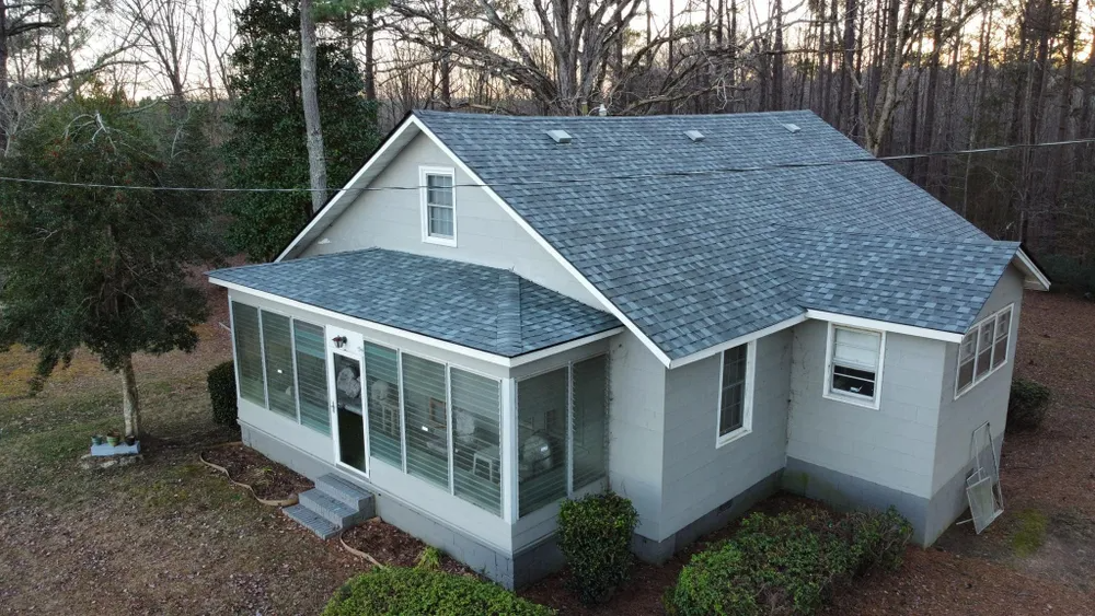 Small gray house with a screened front porch, white trim, and a blue roof in a wooded yard at dusk