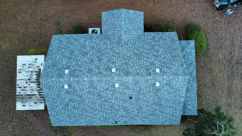 Top-down aerial view of a blue-gray house roof with chimneys, driveway, and surrounding yard.