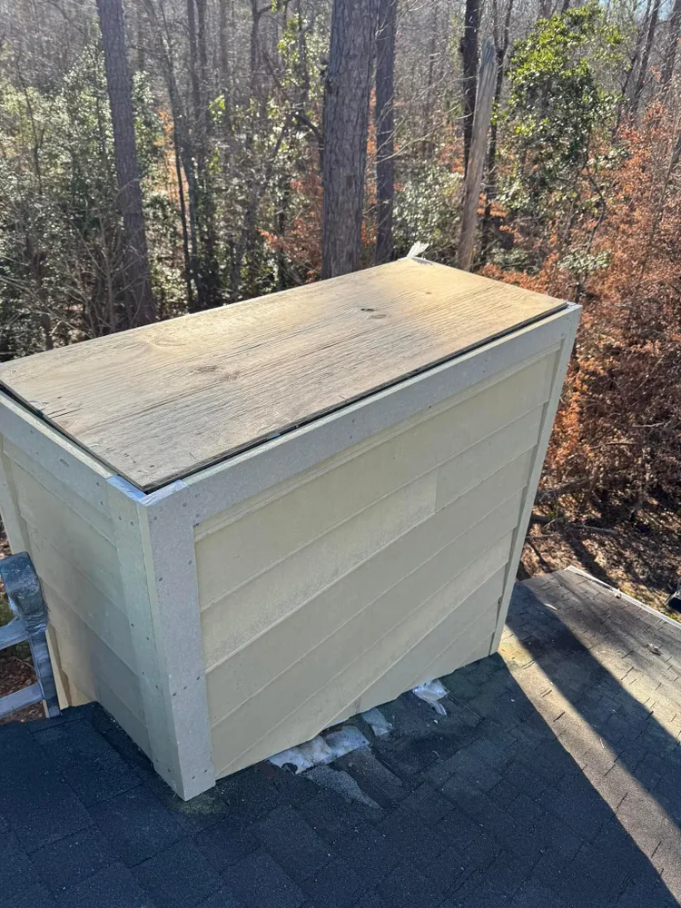 Beige rooftop storage shed beside trees, with a flat tan lid on a shingled roof.