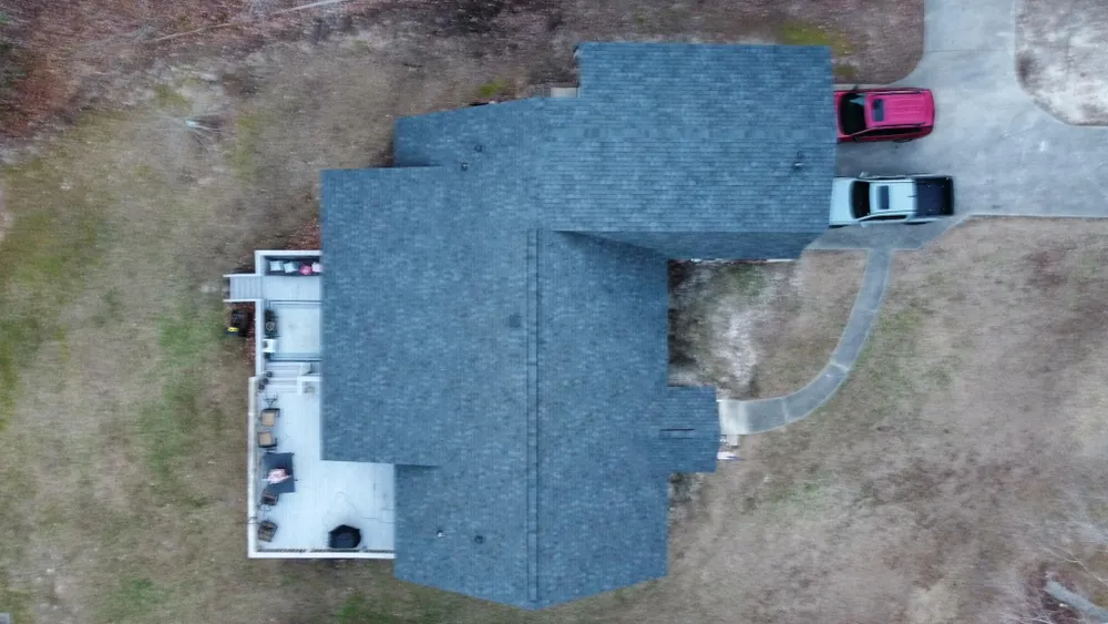 Aerial view of a large gray-roofed house with two parked cars and a curved driveway on a bare yard.