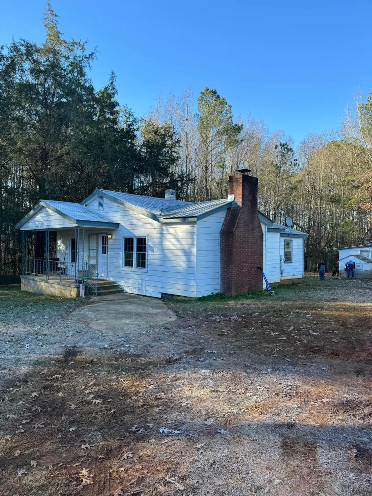 White farmhouse with red brick chimney in a wooded yard under a clear blue sky.