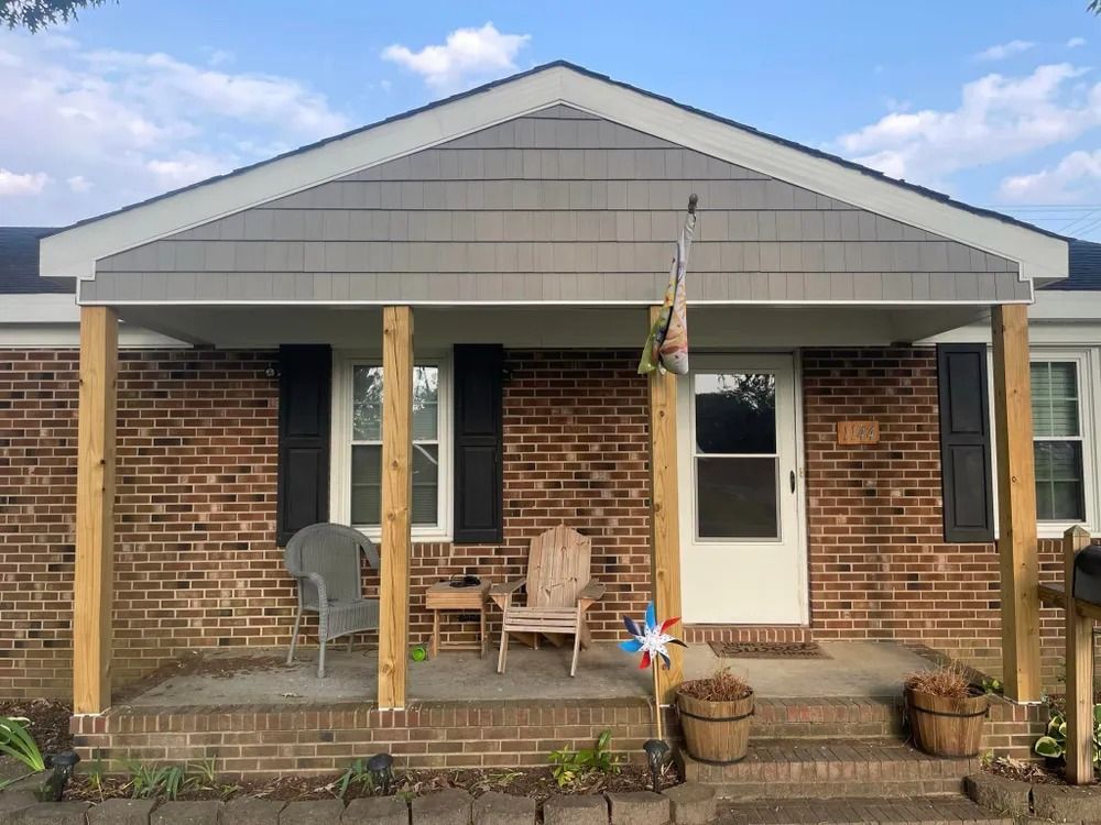 Brick house front porch with white trim, chairs, and potted plants under a gabled roof.