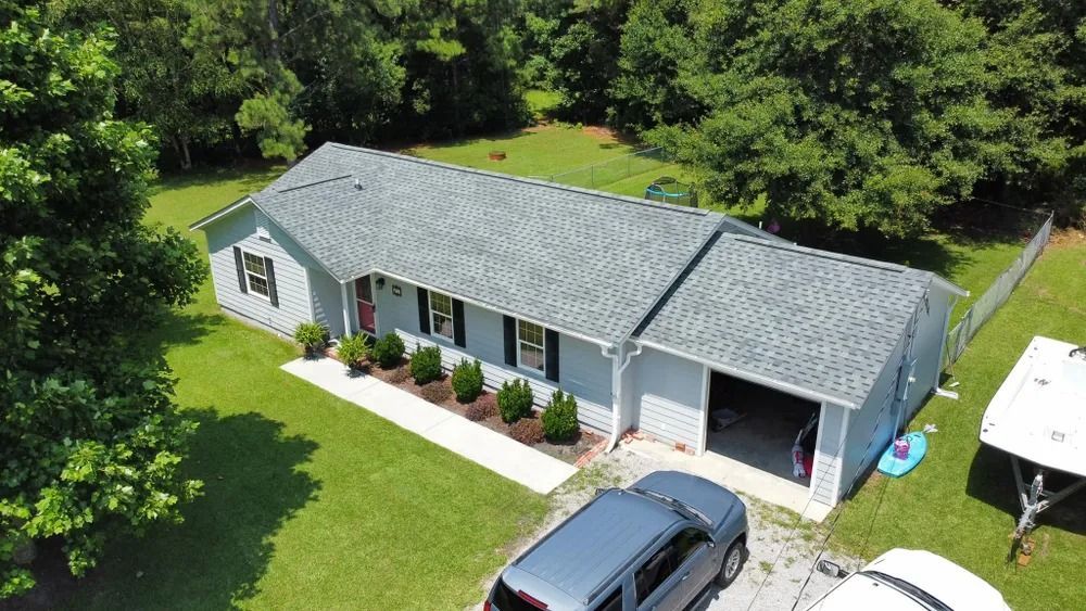 Aerial view of a single-story gray house with a driveway, lawn, and trees surrounding it