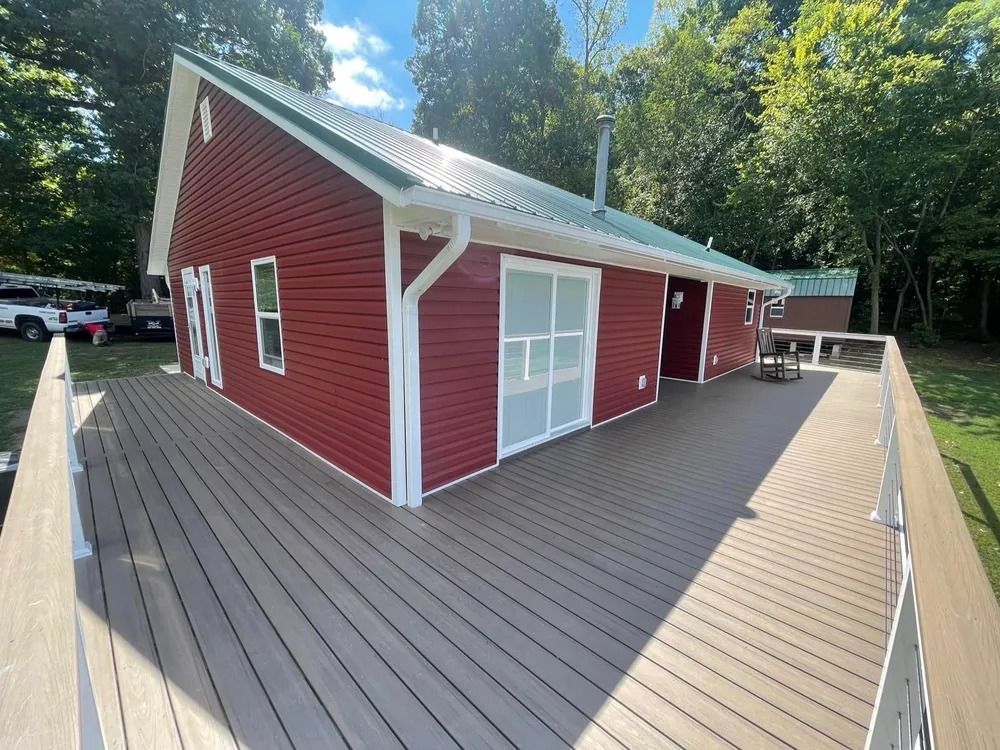 Red outbuilding with white trim on a wooden deck, surrounded by trees under a sunny sky.