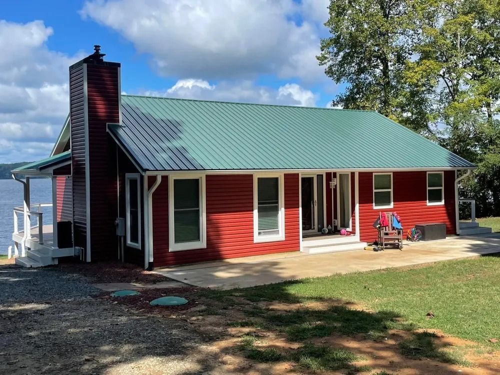Red cabin with green metal roof and porch, set on a grassy lakeside under a partly cloudy sky