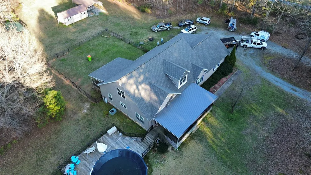 Aerial view of a gray house with a dark roof, driveway, parked cars, and a trampoline in the yard.