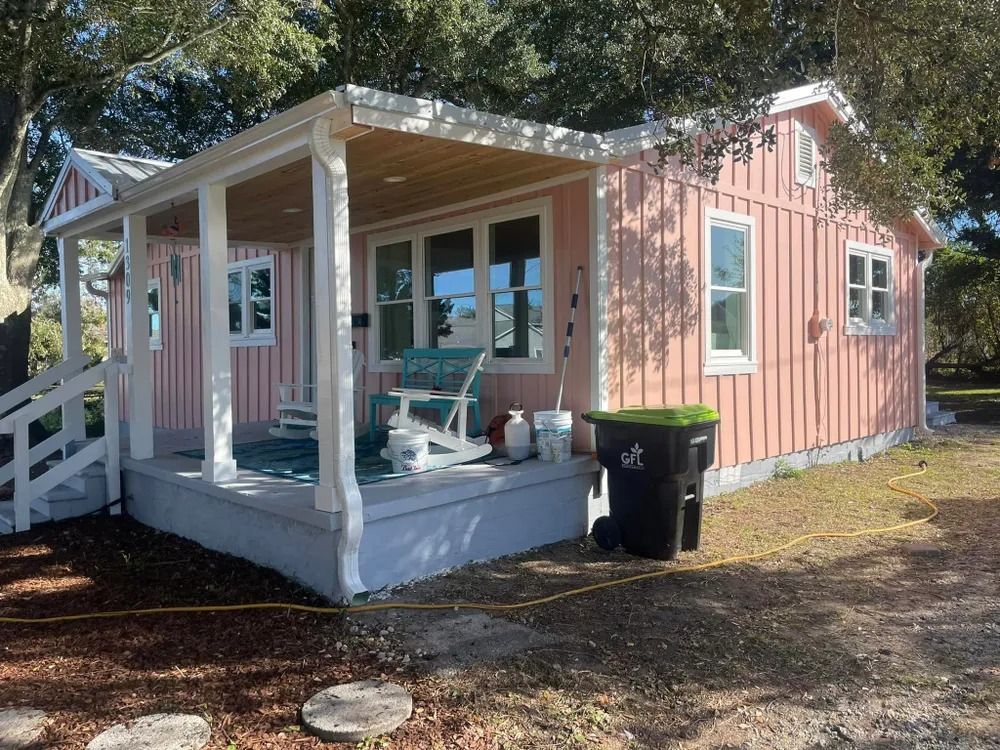 Small pink cottage with white porch, teal chairs, and a black trash bin in a sunny yard