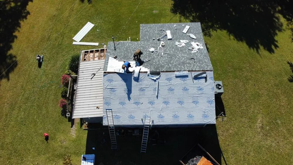 Aerial view of workers on a blue-gray roof in a grassy yard, with ladders and construction materials.