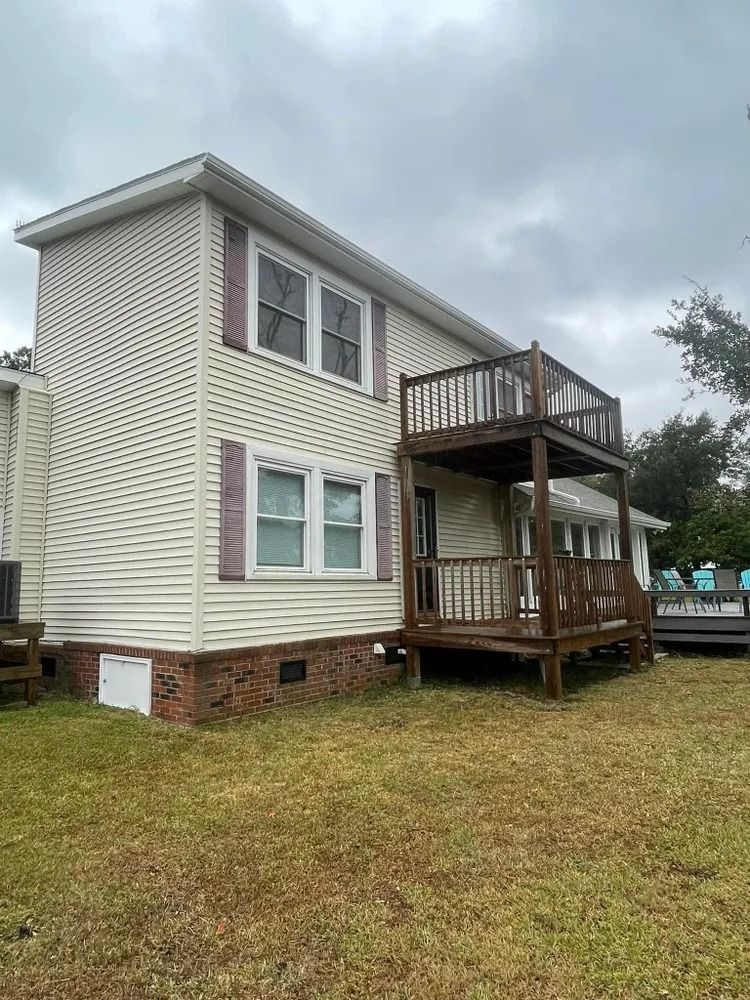 Two-story beige house with a wooden porch and balcony on a grassy lot under cloudy skies