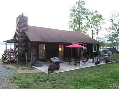 Rustic log cabin with a red umbrella on the patio, set in a grassy yard near trees