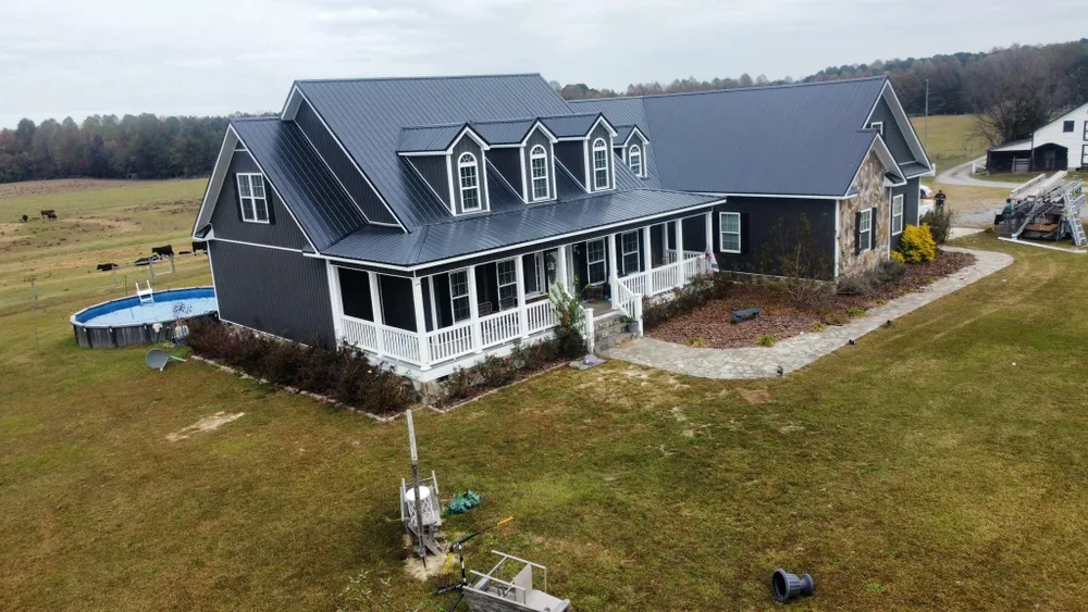 Large gray house with white porch, dark roof, and dormer windows on a rural lot under overcast skies