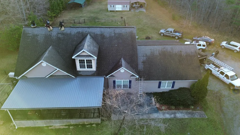 Aerial view of a house with a metal-roofed porch, surrounded by trees and parked vehicles.