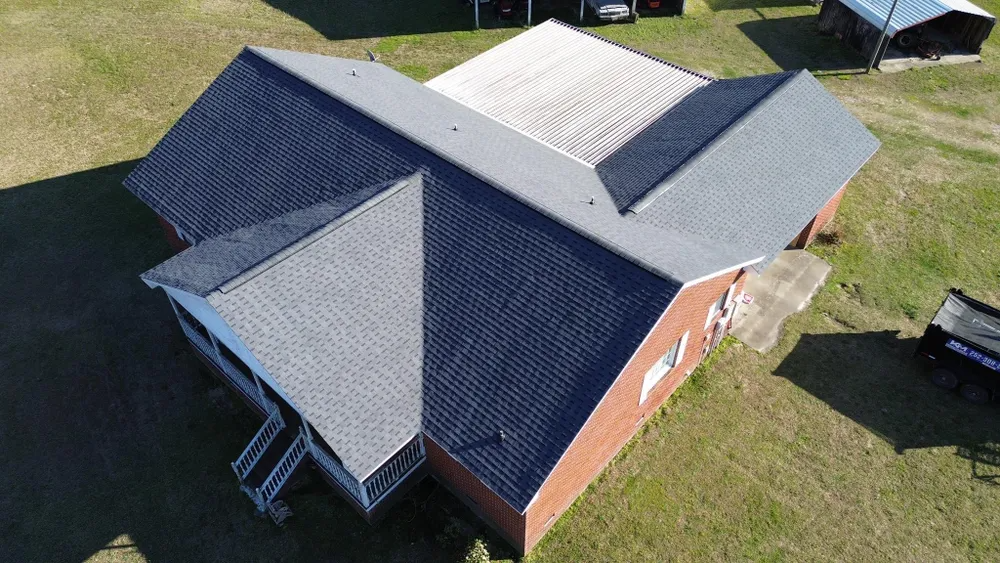 Aerial view of a brick house with a dark gray roof and white roof sections on a grassy lot.