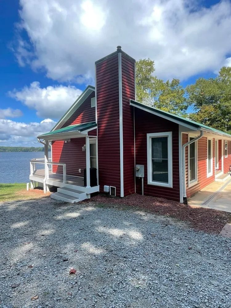 Red lakeside cabin with white trim and brick chimney beside a gravel drive under a blue sky