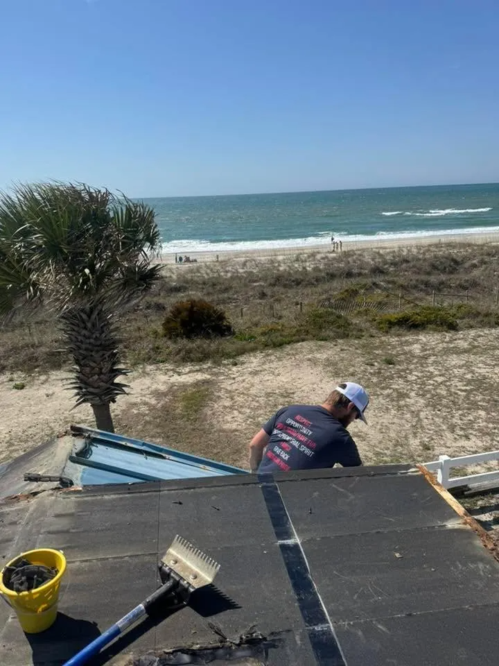 Person on a roof repairing shingles near the beach, with ocean and sky in the background.