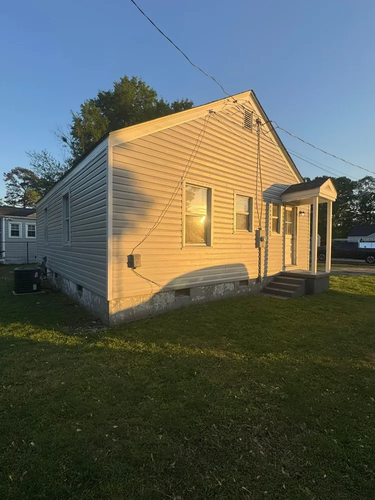 Small beige house with sunlit siding, a front porch, and a grassy yard at sunset