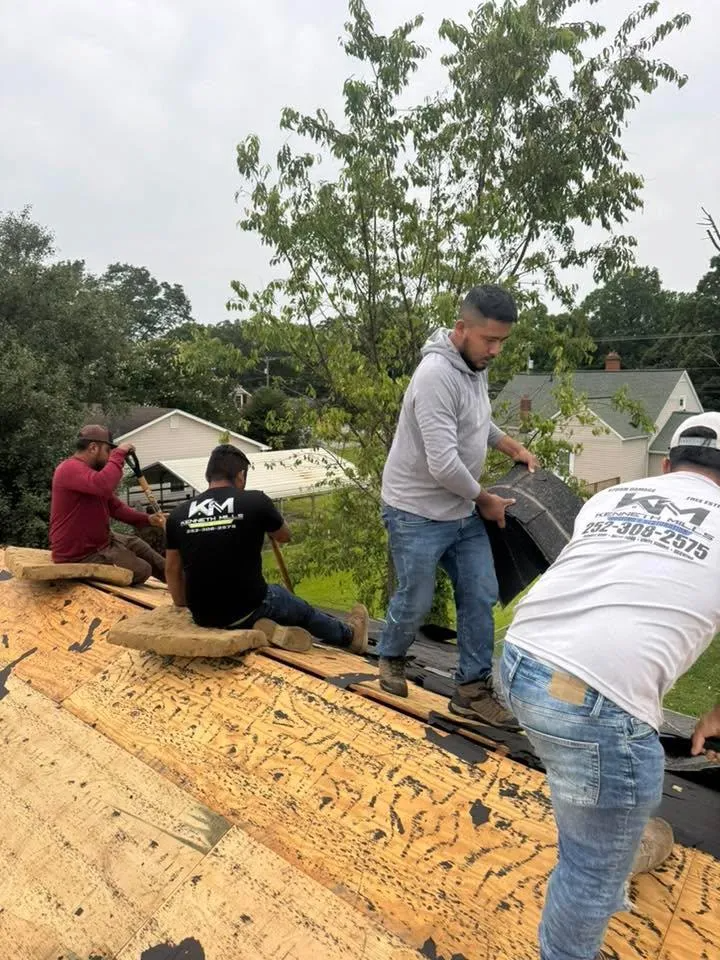 Workers installing shingles on a roof with trees and houses in the background