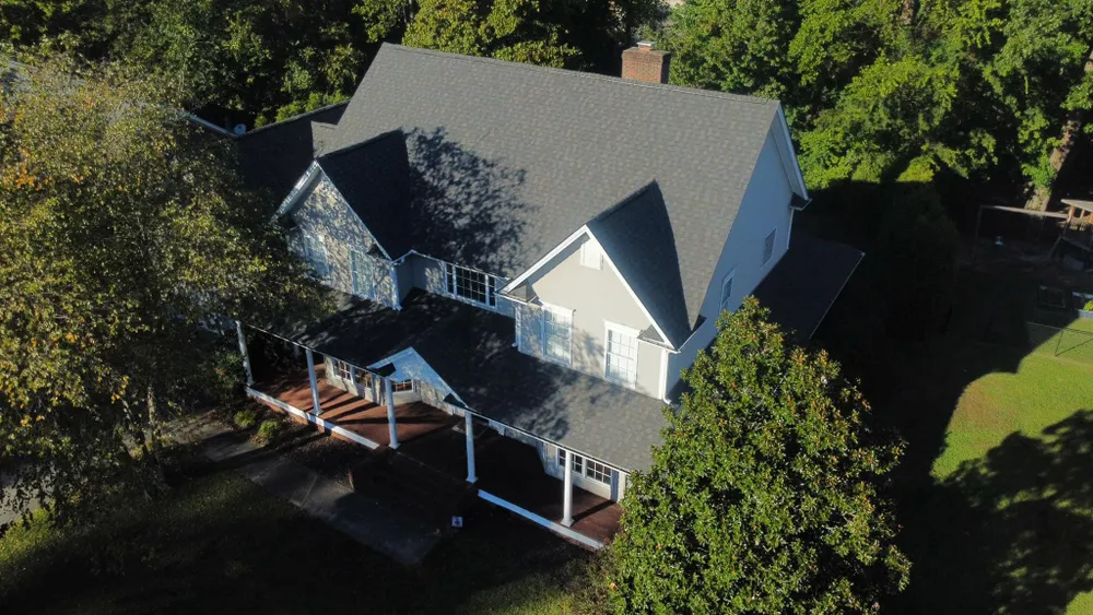 Aerial view of a gray-roofed house with a front porch, surrounded by trees and yard shadows.