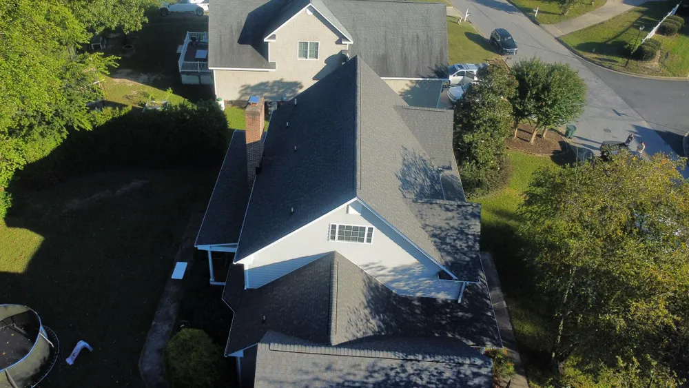 Aerial view of a gray house with a dark roof, driveway, and green trees in a residential neighborhood