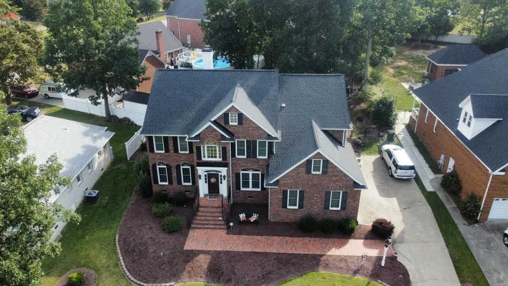 Aerial view of a brick suburban house with a dark gray roof, front porch, and landscaped yard.