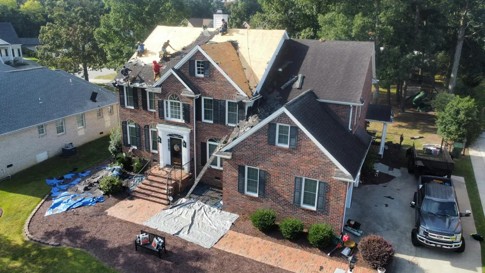 Aerial view of a brick house with white trim, a driveway, parked cars, and blue tarps in the yard.
