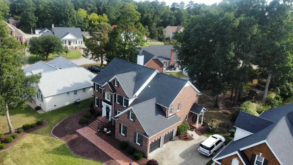 Aerial view of a brick suburban house with a dark roof, driveway, and tree-lined yard.