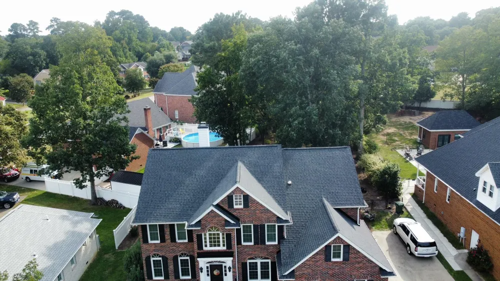 Aerial view of a brick suburban house with a dark roof, driveway, and trees surrounding it