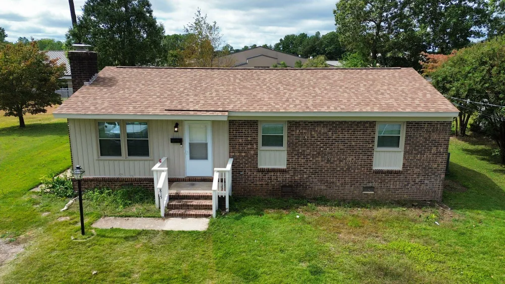 Small brick house with tan roof, white porch, and green lawn under a cloudy sky