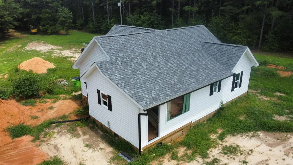 White house with gray roof under construction, surrounded by dirt and trees.