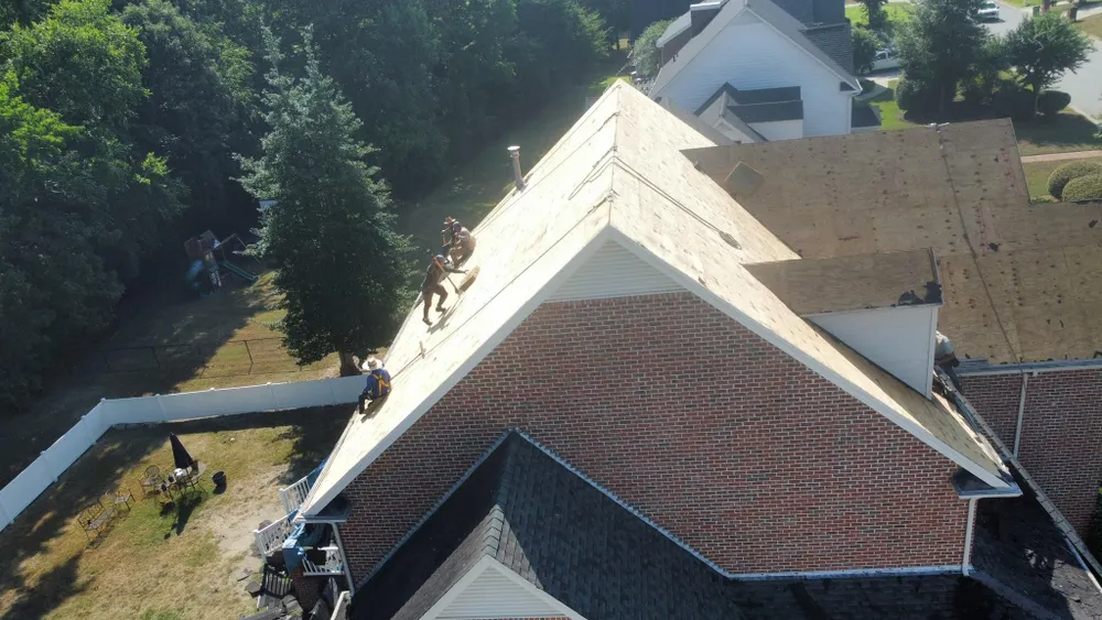 Roofers working on a house roof with exposed plywood and shingles; white gutter visible along the edge.