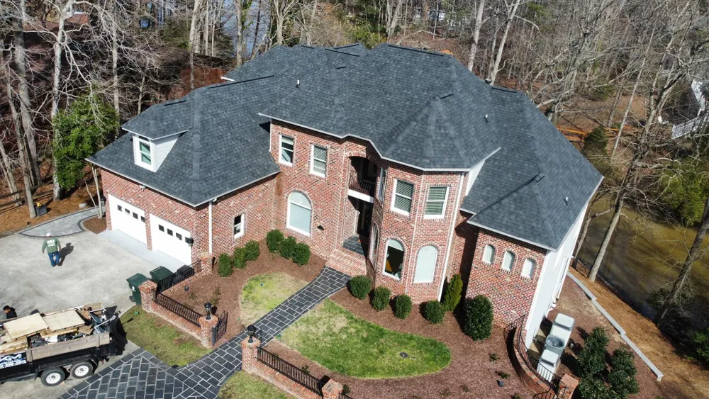 Aerial view of a large brick house with a dark roof, driveway, and wooded yard.