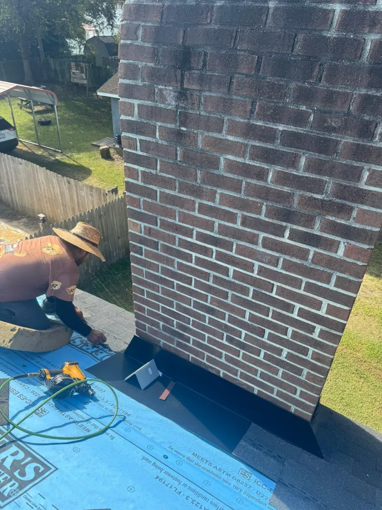 Worker repairing shingles on a brick chimney roof with blue underlayment.