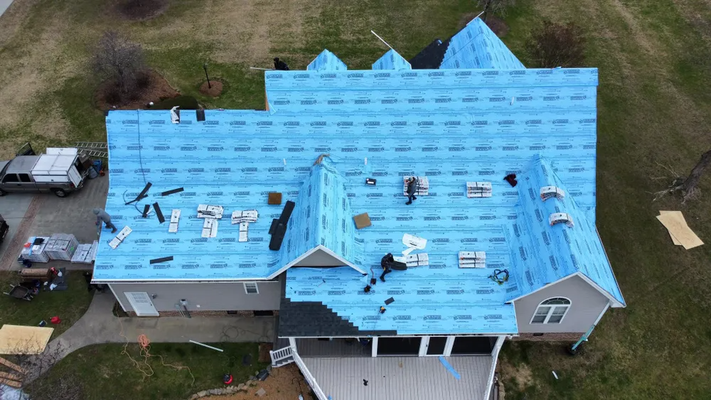 Aerial view of a house roof under construction, covered with blue underlayment and scattered tools and materials.