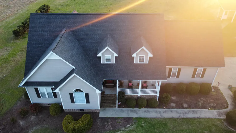 Aerial view of a gray suburban house with white trim and a dark roof in warm sunlight