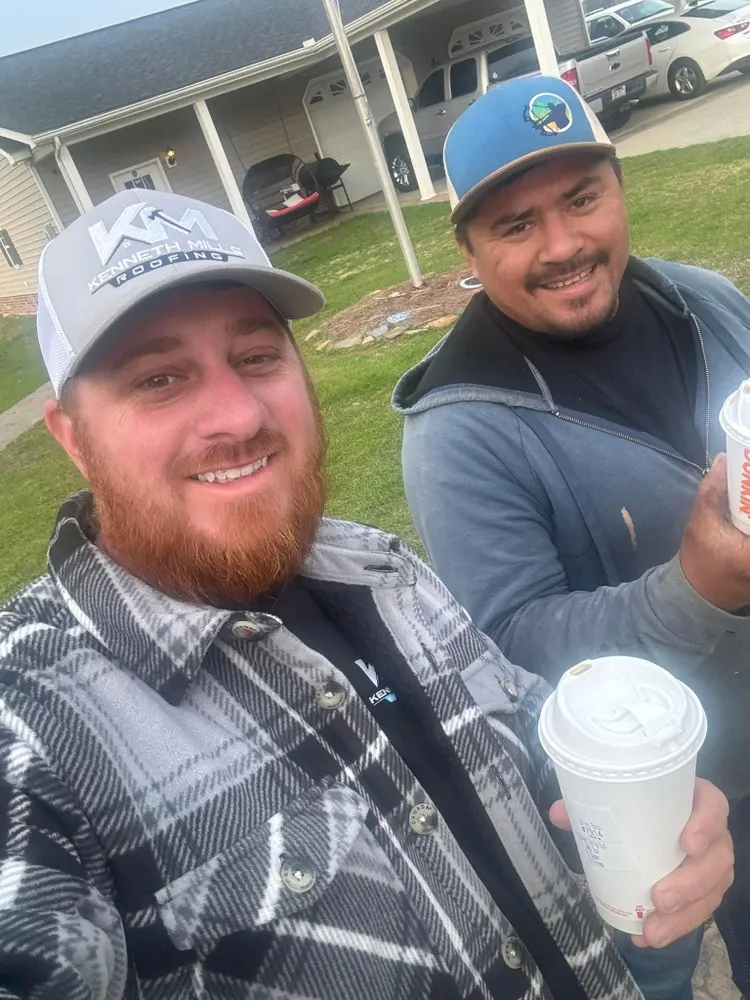 Two smiling men outdoors holding drinks, one in a plaid shirt and cap, the other in a blue cap and jacket.