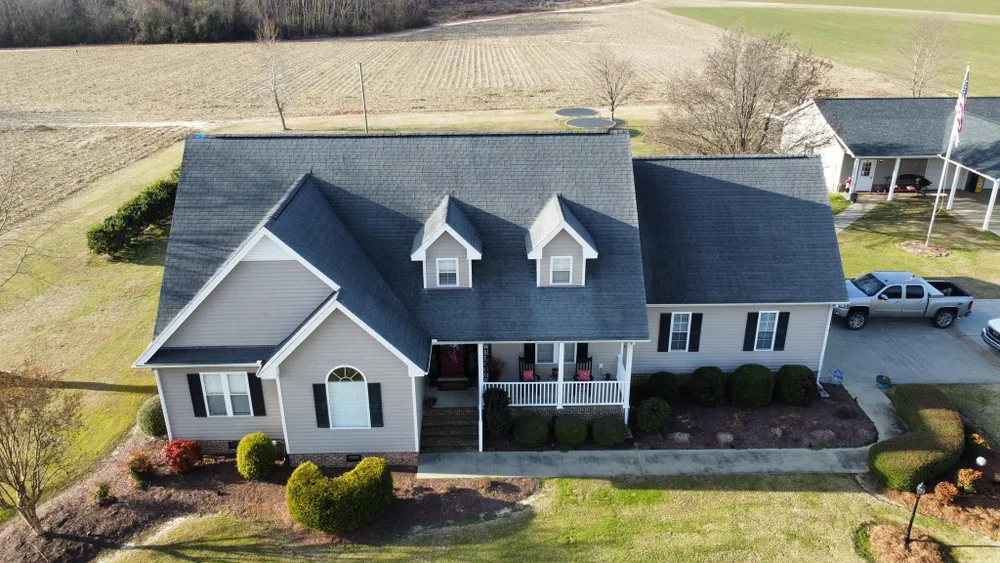 Aerial view of a gray suburban house with a front porch, dormer windows, and surrounding fields.