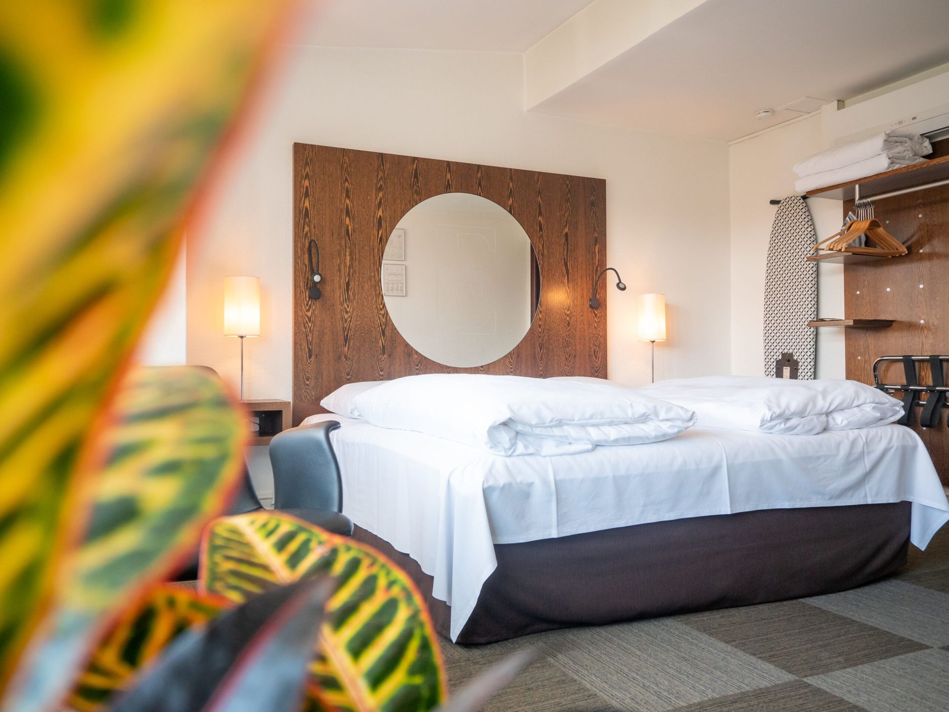 Bedroom with a large bed, round mirror, and wooden headboard. A plant is in the foreground.