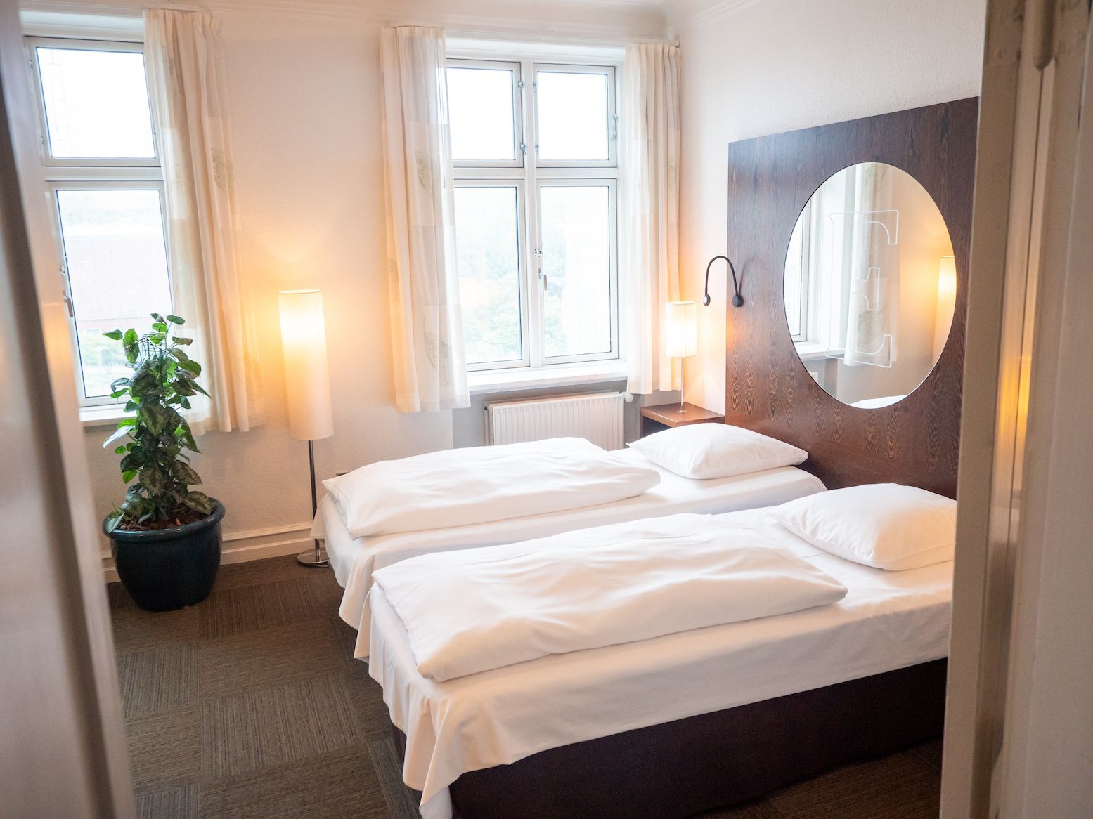 Et lyst hotelværelse med to hvidredte senge, et stort rundt spejl på en hovedgærde af træ, to vinduer og en potteplante.