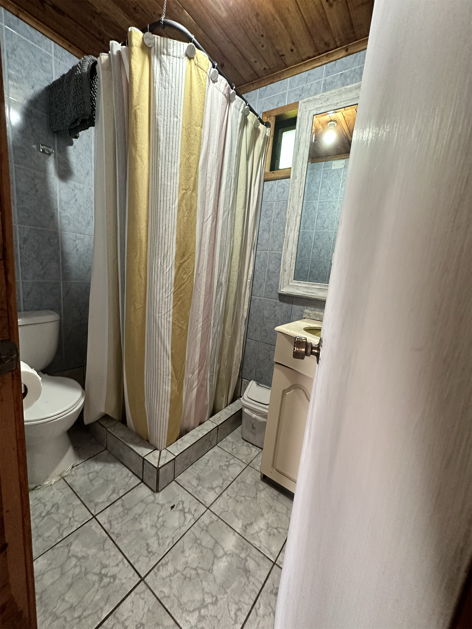 Baño pequeño con ducha, inodoro y lavabo. Cortina de ducha beige y amarilla. Paredes y suelo de azulejos azules.