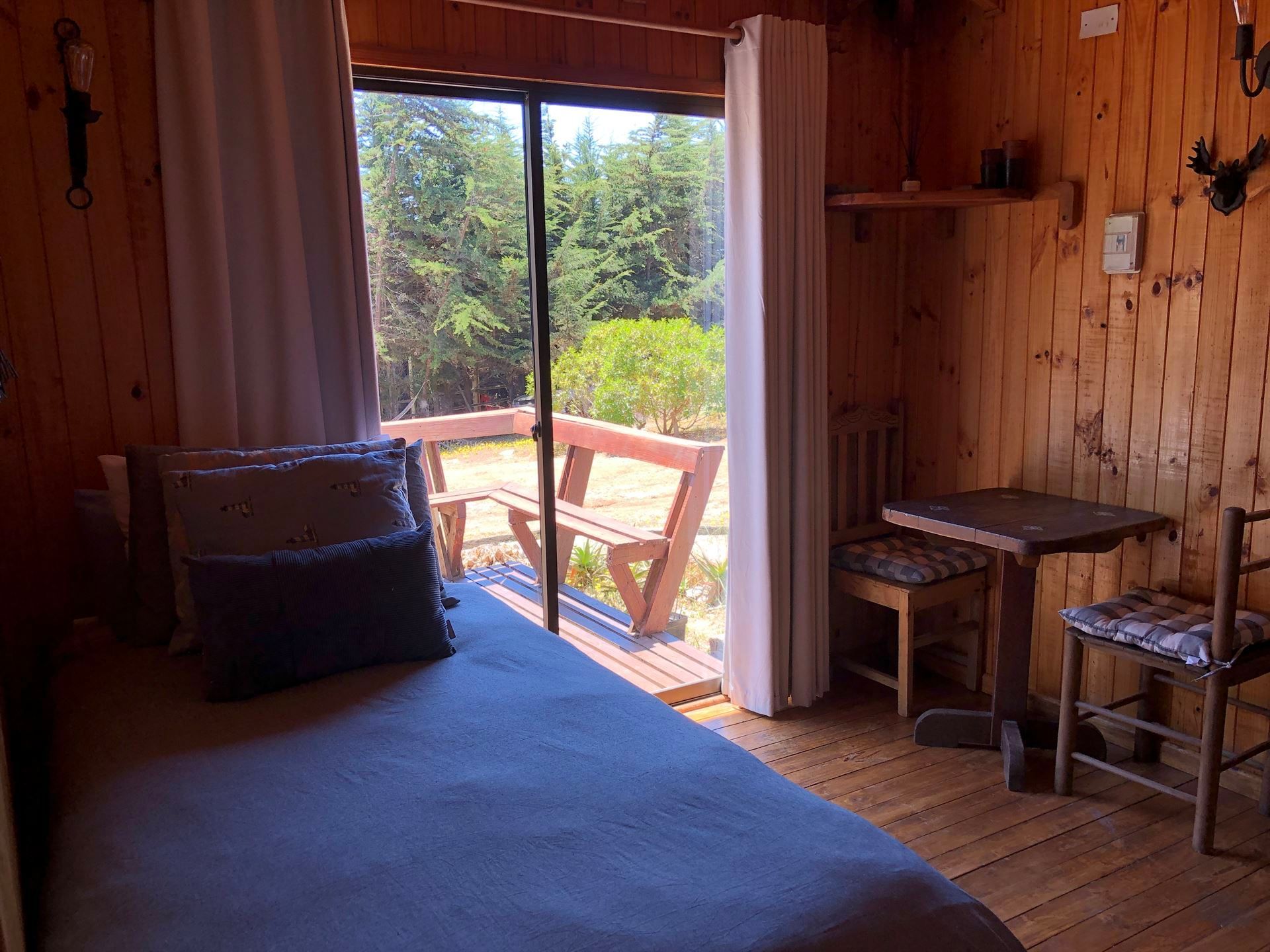 Interior de cabaña de madera con cama, mesa pequeña y puerta corrediza abierta a banco exterior.
