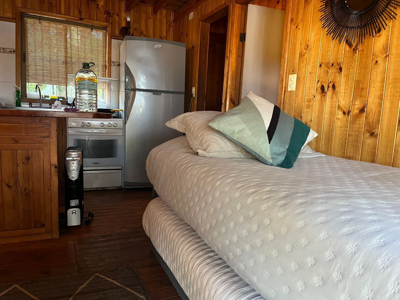 Interior de cabaña pequeña con cama, refrigerador y cocina. Paredes de madera y luz natural.