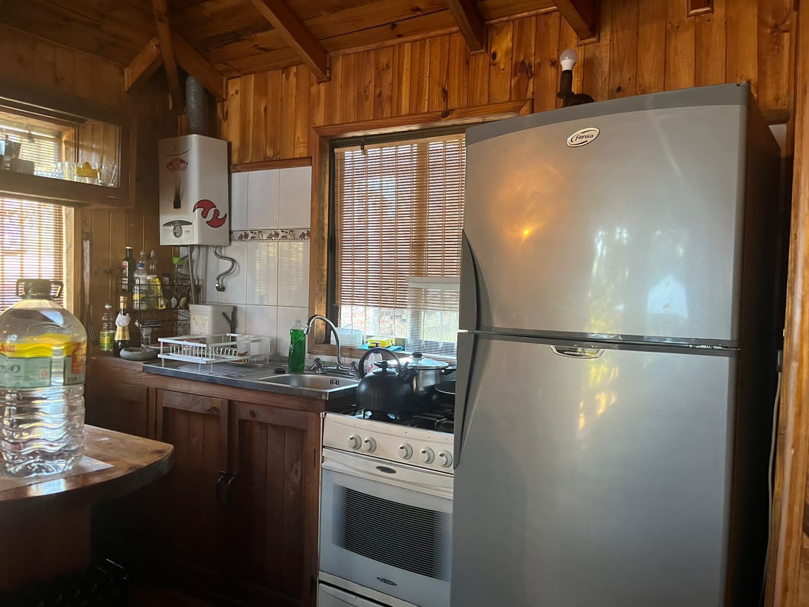 Pequeña cocina tipo cabaña con paredes de madera, estufa, refrigerador y ventana con persianas.