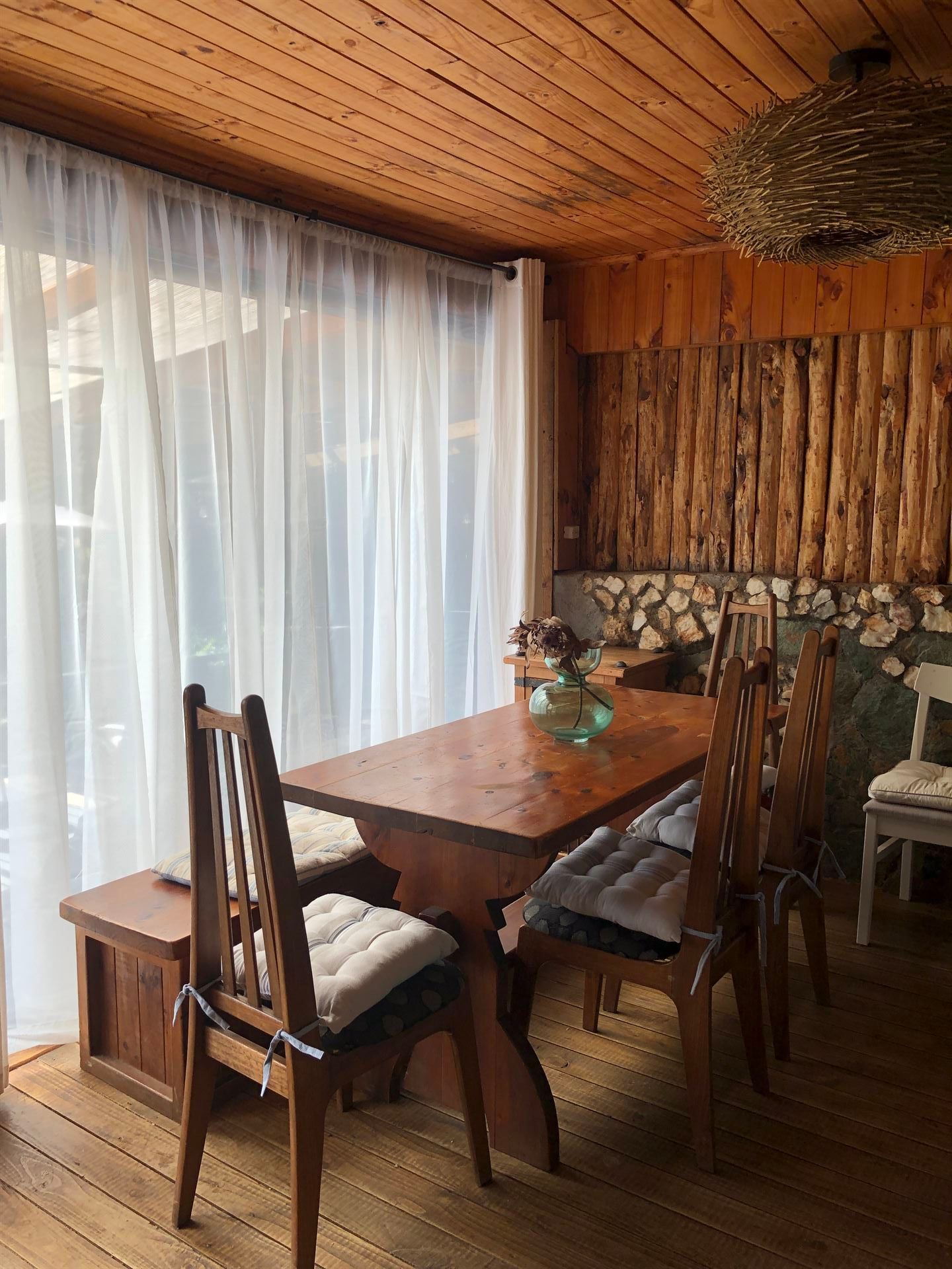 Mesa de comedor y sillas de madera en una habitación rústica de madera con cortinas blancas transparentes.