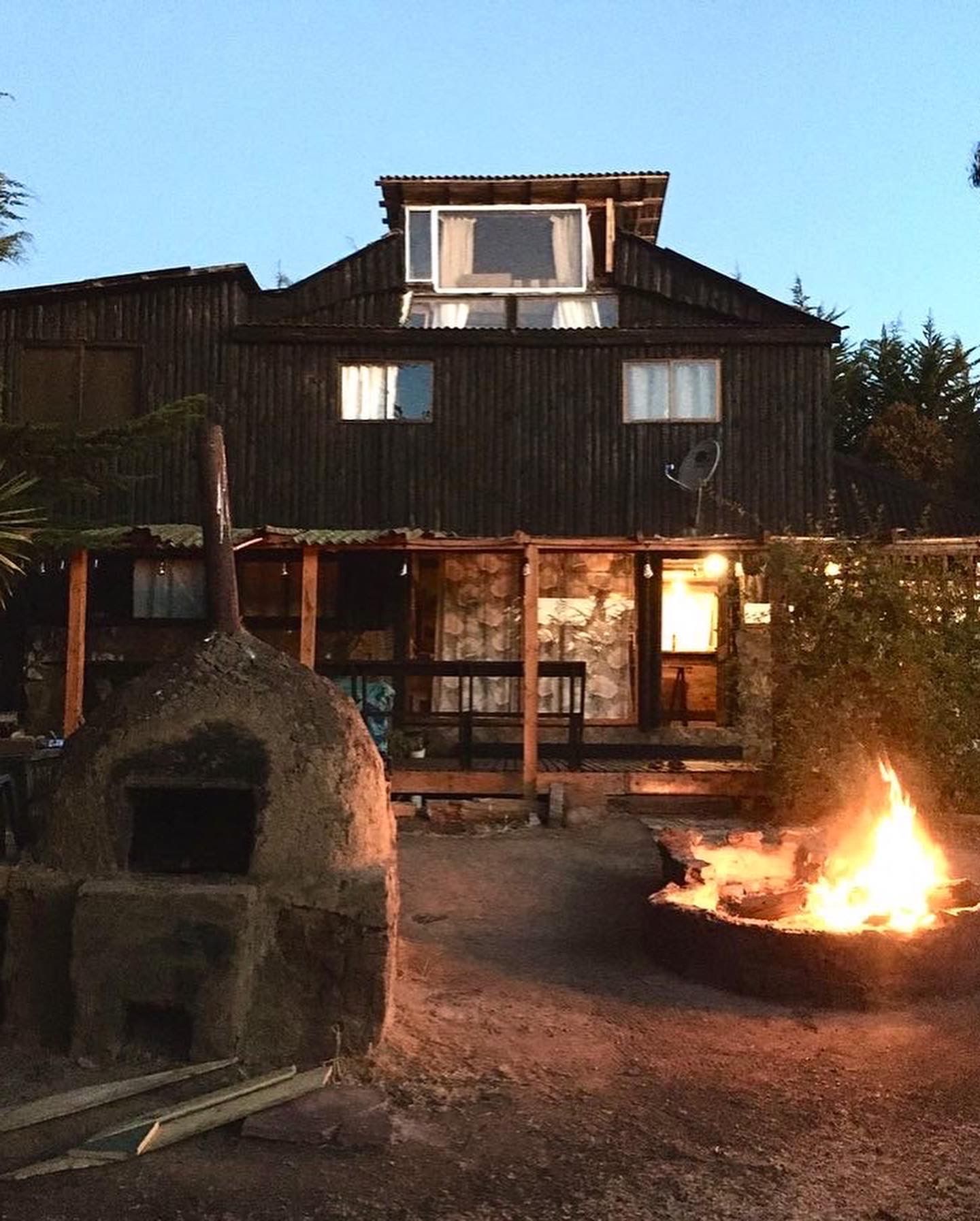 Casa de madera con horno exterior y fogata al anochecer.