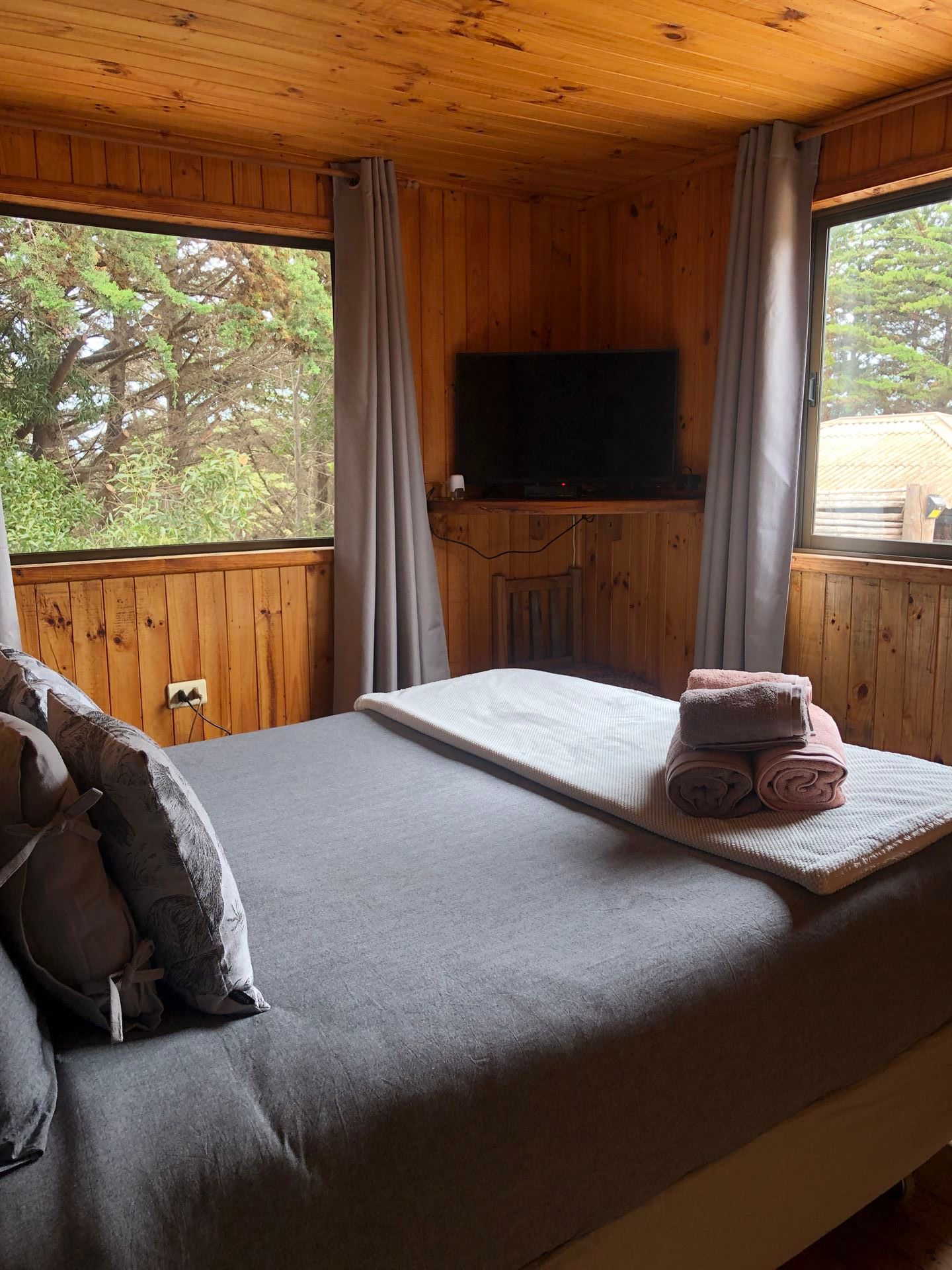 Dormitorio con paredes y techo de madera, cama gris, TV y dos ventanas con vista a los árboles.