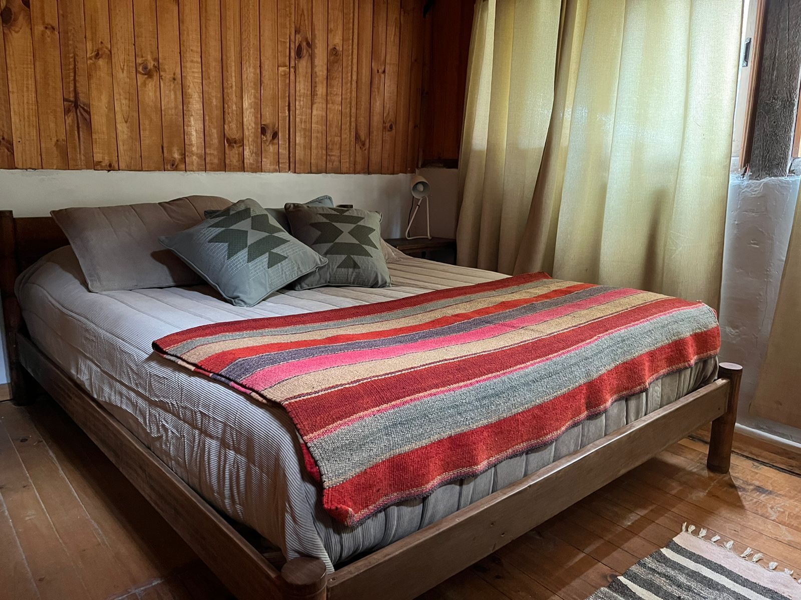Dormitorio con paneles de madera, una cama con manta de colores, dos almohadas y una ventana con cortinas transparentes.