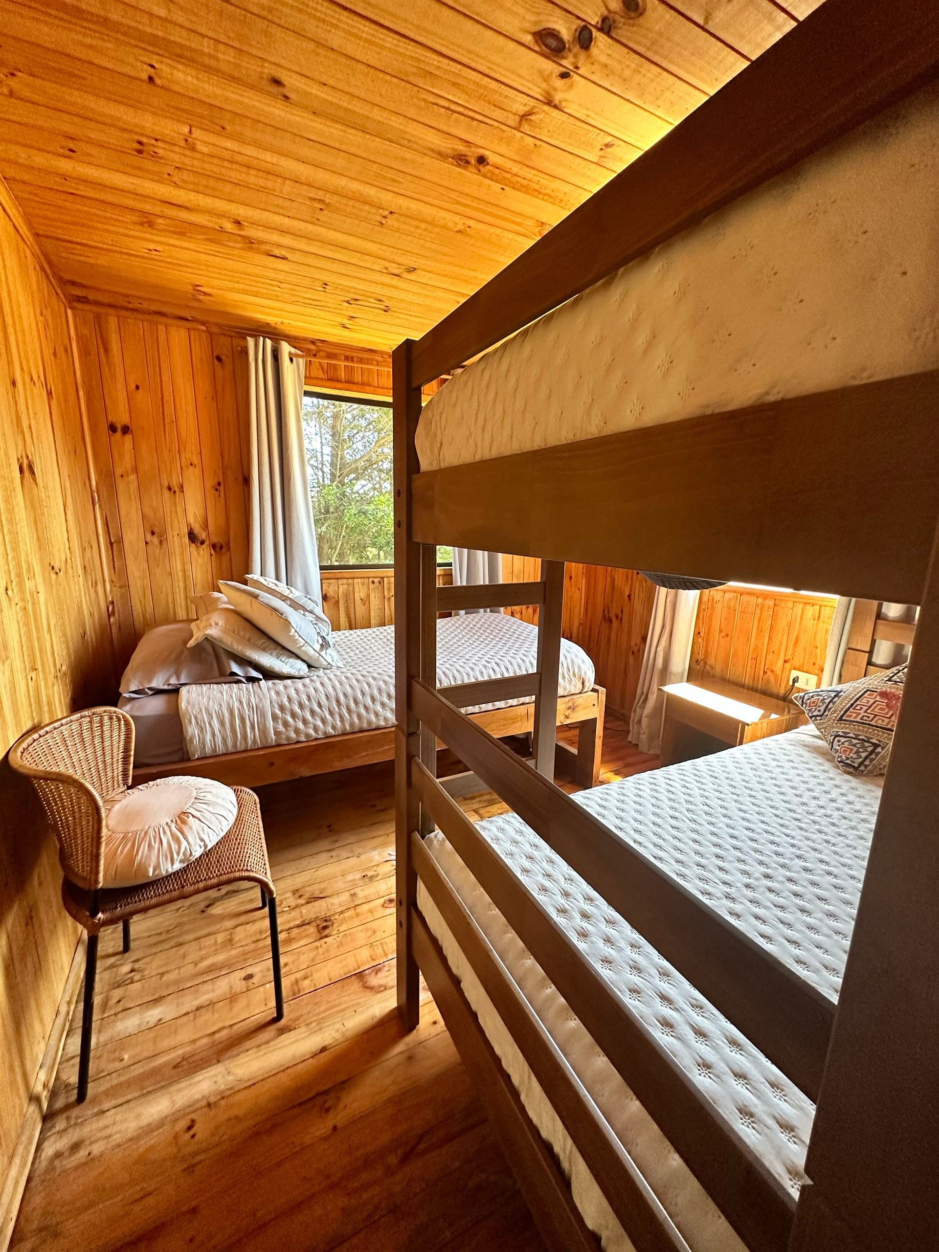 Literas de madera y una cama individual en una habitación revestida de madera con ventana y silla de mimbre.